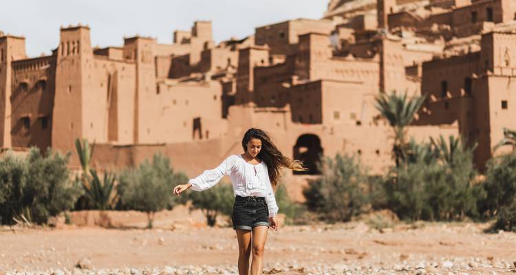 Mujer caminando frente al antiguo sitio de Ait Benhaddou con palmeras y edificios de piedra.