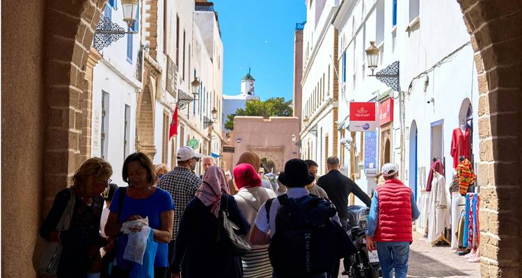 Des foules parcourent les boutiques et cafés le long d'une rue étroite ensoleillée encadrée par des arches de pierre dans la médina d'Essaouira.