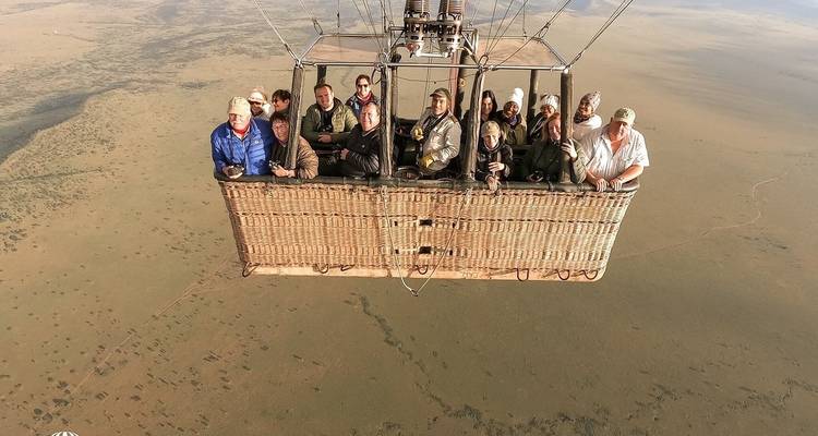 Een groep mensen in een luchtballonmandje die over een uitgestrekte savanne vliegt.