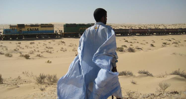 Homme en tenue traditionnelle face à un train dans le désert.