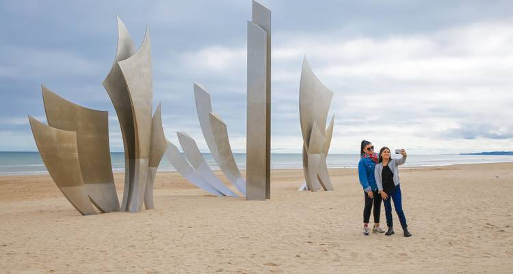 Dos mujeres se toman una selfie junto a la escultura Les Braves del Día D en Playa Omaha bajo un cielo gris