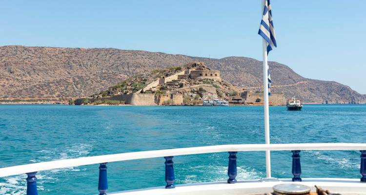 Aussicht vom Boot auf die Festung der Insel Spinalonga im Voraus, während die griechische Flagge über dem leuchtend blauen Meer weht.