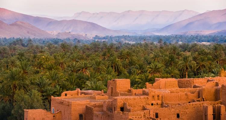 Sonnenbeschienene Ruinen einer Adobe-Kasbah überblicken eine weite Dattelpalmen-Oase vor dem Hintergrund diesiger Bergketten.