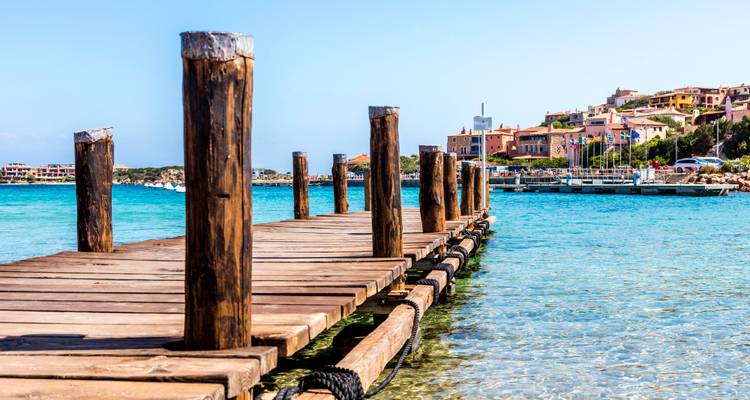 Jetée en bois s'étendant sur une eau turquoise cristalline vers une marina et des bâtiments aux teintes pastel.