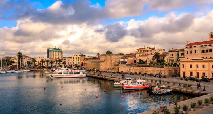 Port d'Alghero avec des bateaux amarrés, des murailles médiévales et des maisons de ville aux couleurs vives sous un ciel aux nuages spectaculaires.