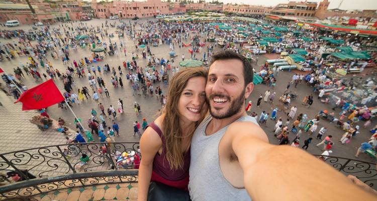 Ein Paar, das ein Selfie mit einem belebten Marktplatz im Hintergrund macht.