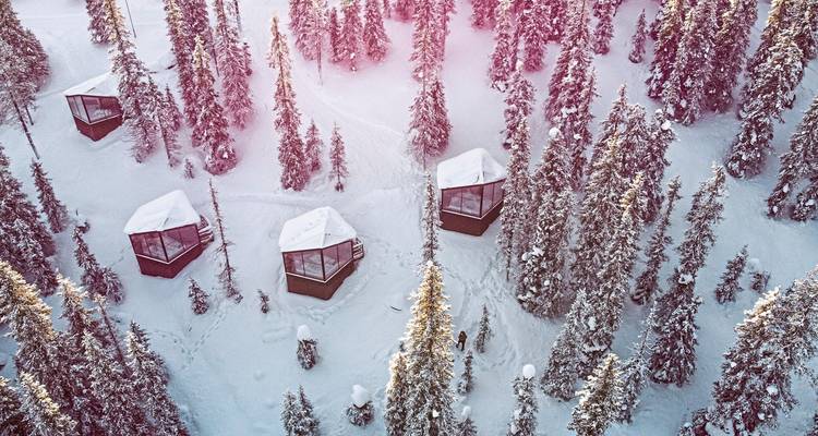 Vue aérienne de cabanes enneigées au milieu d'un terrain forestier dense, baignées de soleil.