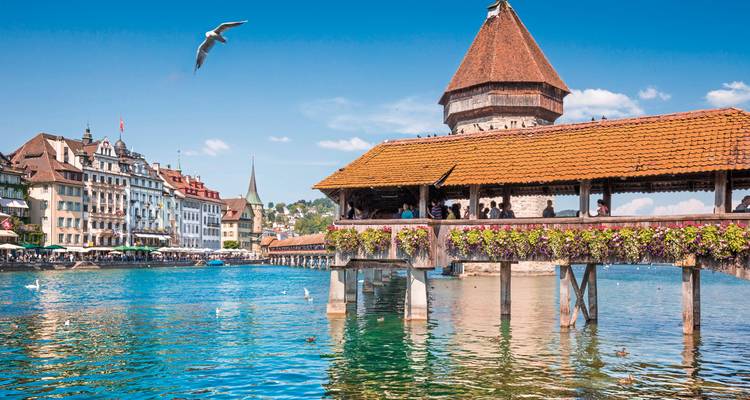 El histórico Puente de la Capilla de madera se extiende sobre el azul río Reuss con jardineras de flores, cisnes y el casco antiguo de Lucerna al fondo.