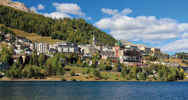 Elegantes hoteles y edificios de St. Moritz se alzan sobre un tranquilo lago alpino enmarcado por un bosque de coníferas.