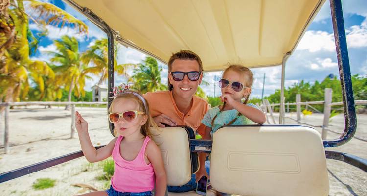 Famille se déplaçant en voiturette de golf sur une plage ensoleillée avec des palmiers en arrière-plan.