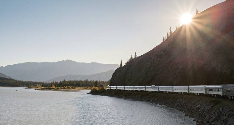 Zug, der bei Sonnenaufgang neben einem großen See mit Bergkulisse fährt.