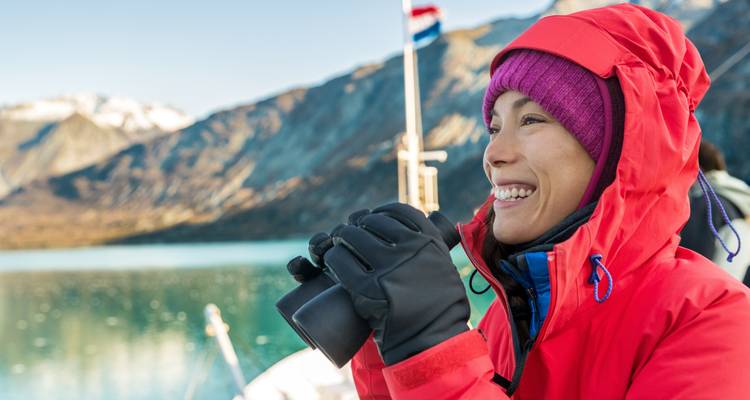 Person in a red jacket holding binoculars on a boat with mountains and water in the background.