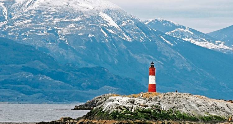 Red and white lighthouse on rocky shores with mountains.