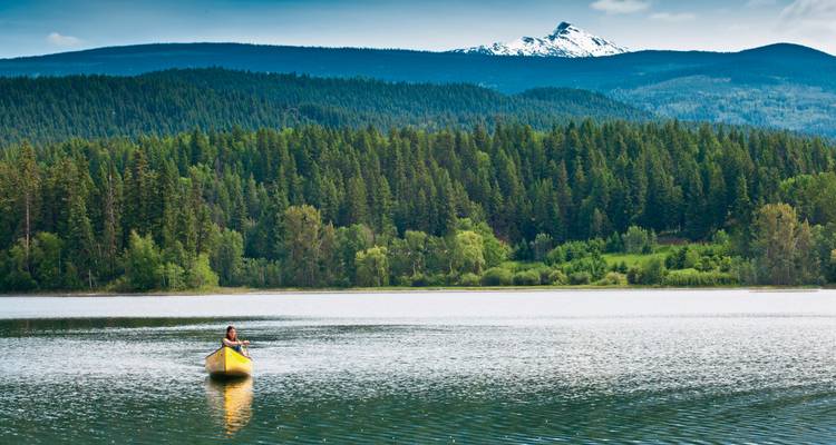 Person, die in einem Kanu auf einem ruhigen See mit einer Bergkette im Hintergrund paddelt.