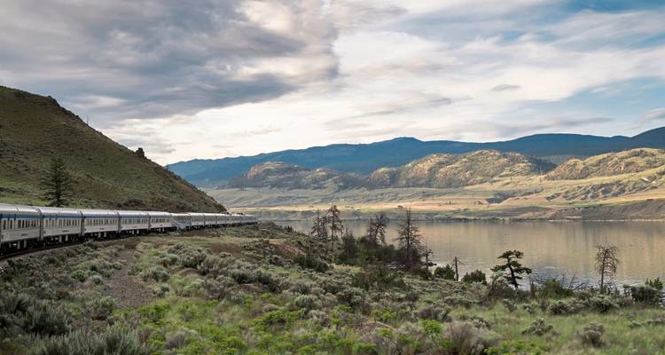 Ein Zug, der an einem See vorbeifährt mit malerischen Bergketten im Hintergrund.