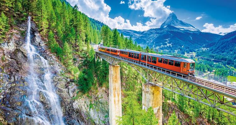 Tren cruzando un puente sobre una cascada en un paisaje montañoso con el Matterhorn en el fondo.