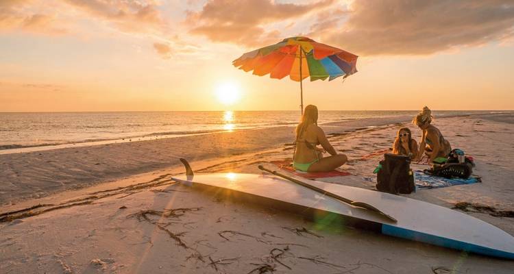 People relaxing on a beach at sunset with a surfboard near the water.