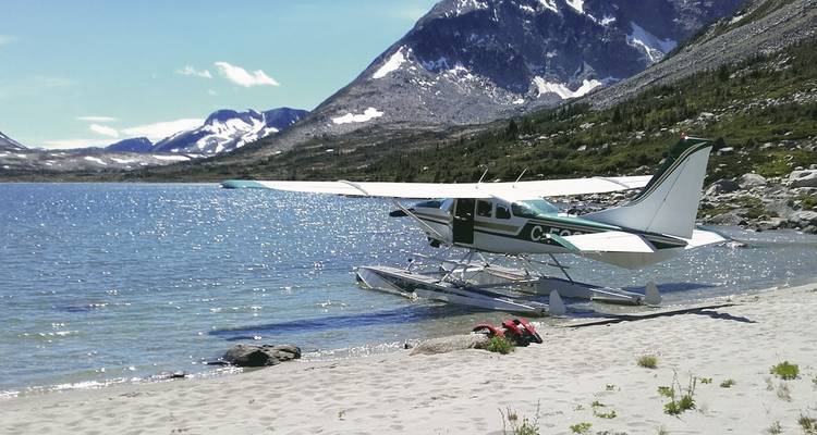 Hidroavión en una orilla arenosa junto a un lago, montañas al fondo.