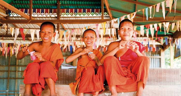 Three young monks laughing and holding items, sitting under a decorated roof.