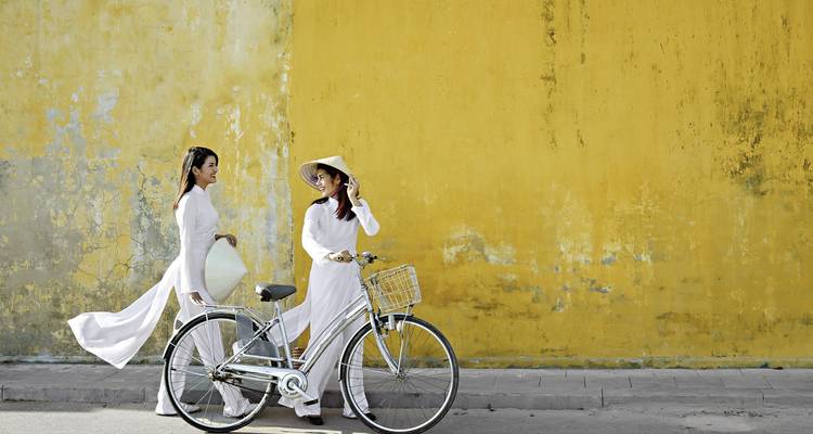 Deux femmes en tenue traditionnelle vietnamienne avec un vélo contre un mur jaune à Hoi An.
