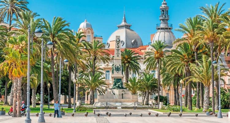 Place avec palmiers, un monument et des bâtiments ornés.