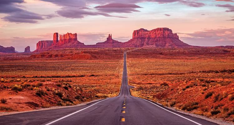 Carretera larga y recta que conduce a formaciones rocosas rojas bajo un cielo de atardecer.