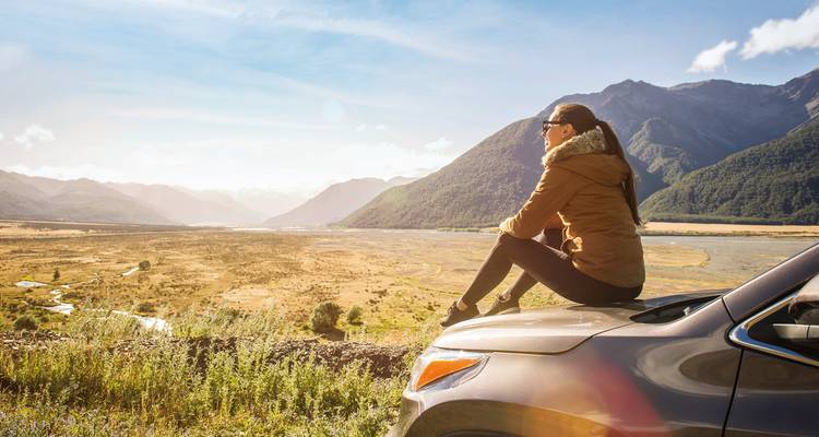 Woman sitting on a car enjoying mountain scenery.