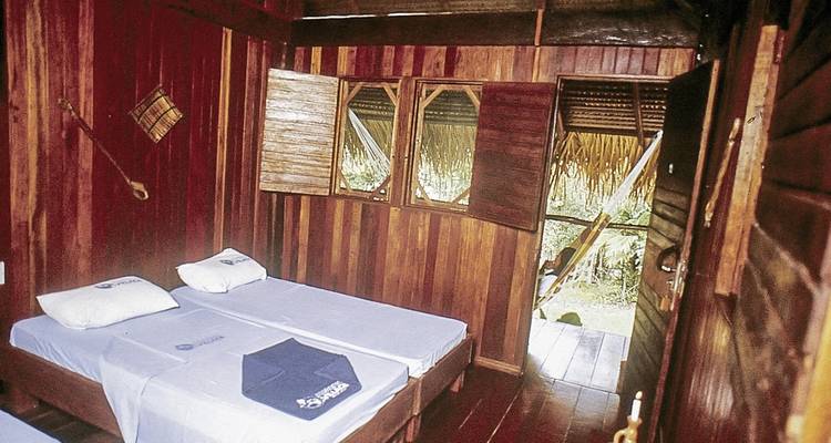 Eenvoudig interieur van een houten hut met twee eenpersoonsbedden en een hangmat bij het raam.