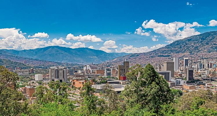 Weite Skyline von Medellín eingebettet in ein grünes Tal, umgeben von Anden-Bergen unter einem leuchtend blauen Himmel.