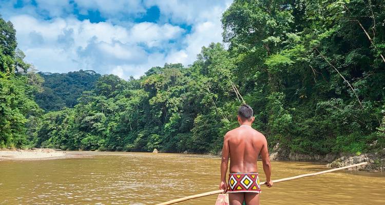 Een persoon in traditionele kleding peddelt op een rivier omringd door dicht regenwoud.