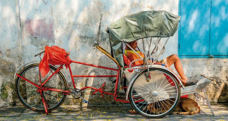 Person resting on a trishaw beside a weathered wall.