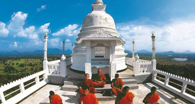 Buddhist monks in red robes meditating at a white hilltop stupa overlooking lush plains