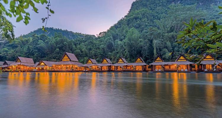 A row of thatched-roof cottages on a river with mountain forest in the background, illuminated at dusk.