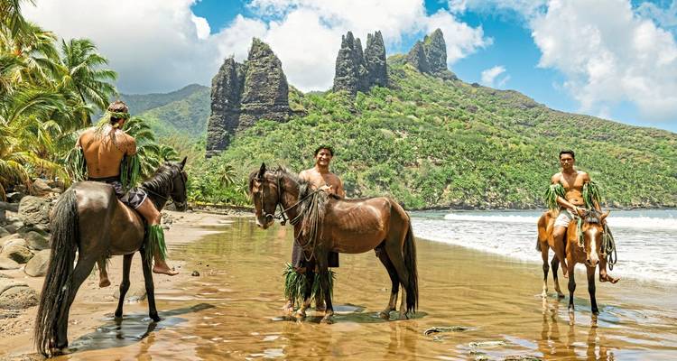 Drie mensen te paard op een tropisch strand met bergen op de achtergrond.