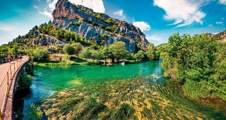 River with lush greenery and rocky hills under a bright blue sky.