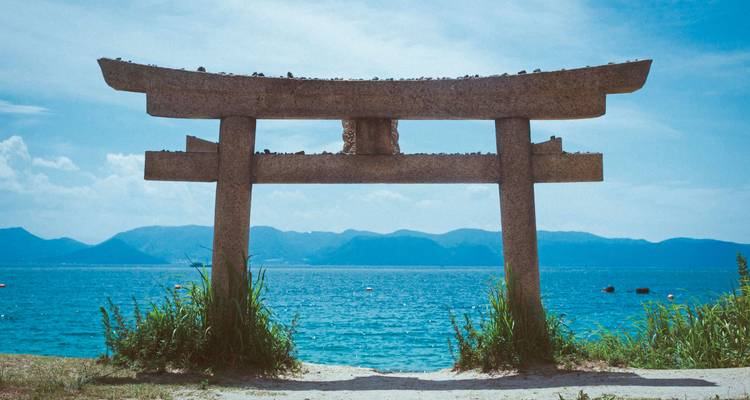 Portail torii en pierre se dressant sur une rive herbeuse dominant un lac bleu tranquille et des montagnes lointaines