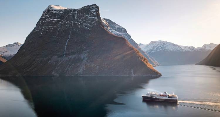 Sunlit fjord with a cruise ship in front of a towering mountain.