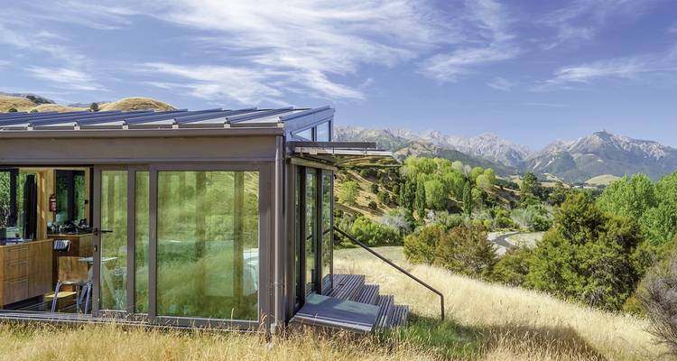 Cabane moderne en verre dans un paysage rural avec des montagnes.