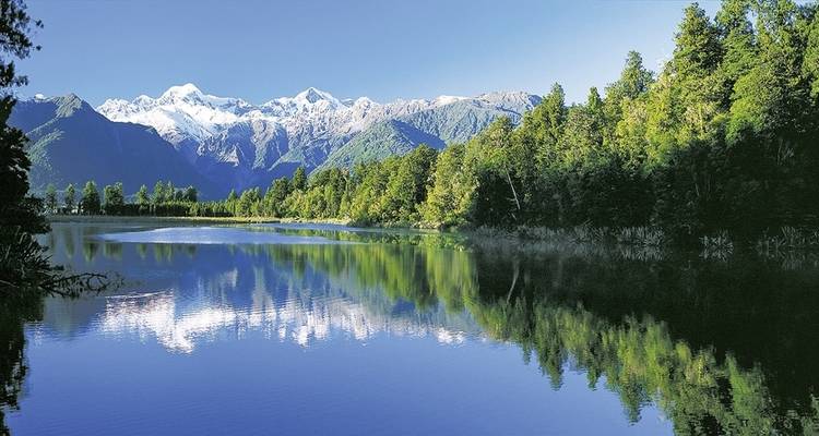 Reflective lake with forested hills and snowy mountains.