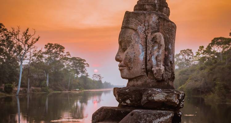 Angkor Thom Gesichtsstatue bei Sonnenuntergang mit Bäumen und einem Fluss im Hintergrund.