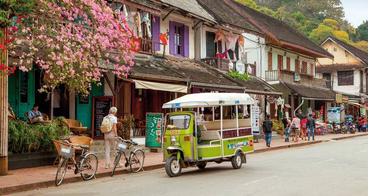 Straßenszene in Luang Prabang mit Tuk-Tuks, Fahrrädern und lebendigen Gebäuden.