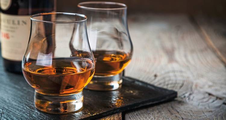 Primer plano de dos vasos de whisky sobre una superficie de madera con una botella al fondo.