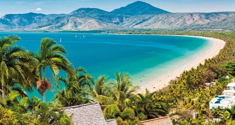 Aerial view of tropical beach with clear blue water and people on the beach, surrounded by lush green vegetation.