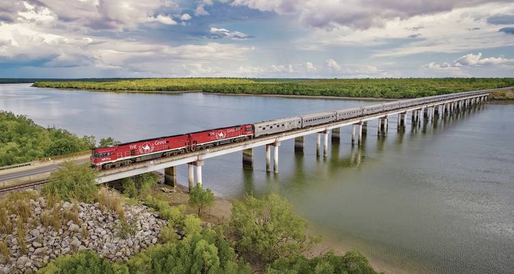 Der Ghan-Zug überquert eine lange Brücke über einen breiten Fluss, der von üppigen grünen Mangrovenwäldern und weitläufigen Himmelsflächen umgeben ist.