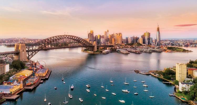 Luftaufnahme des Sydney Harbour mit der Harbour Bridge, die sich über glitzerndes Wasser wölbt, und der Skyline der Stadt, die bei Sonnenuntergang erleuchtet ist.