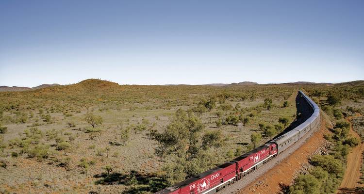 Ein langer roter Zug schlängelt sich durch ausgedehnte australische Outback-Buschlandschaft unter einem klaren blauen Himmel.