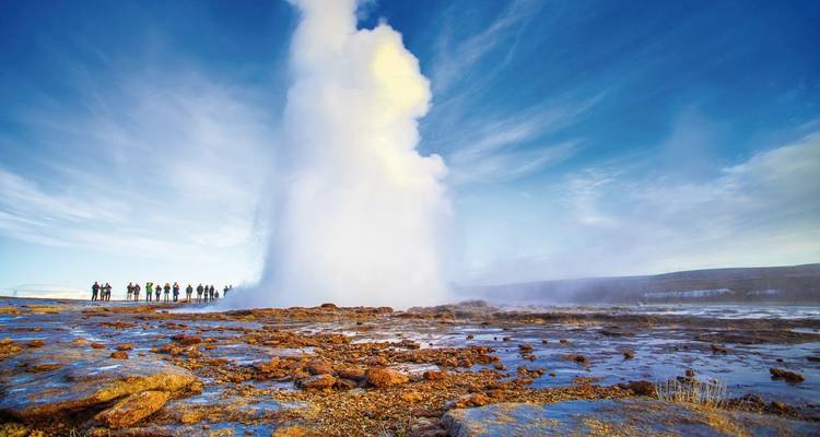 Un énorme nuage de vapeur et d'eau jaillissant d'un geyser tandis que des spectateurs se tiennent debout sur le sol rocheux.