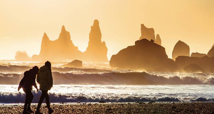 Un couple en silhouette se promène le long d'une plage de sable noir tandis que des formations rocheuses de basalte spectaculaires se dressent contre un ciel de coucher de soleil flamboyant.