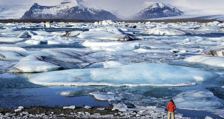 Une personne se tient devant un lagon glaciaire avec des icebergs et des montagnes au loin.