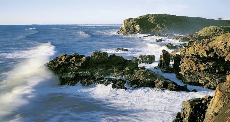 Rochers côtiers escarpés avec des vagues qui s'écrasent contre eux sous un ciel clair.